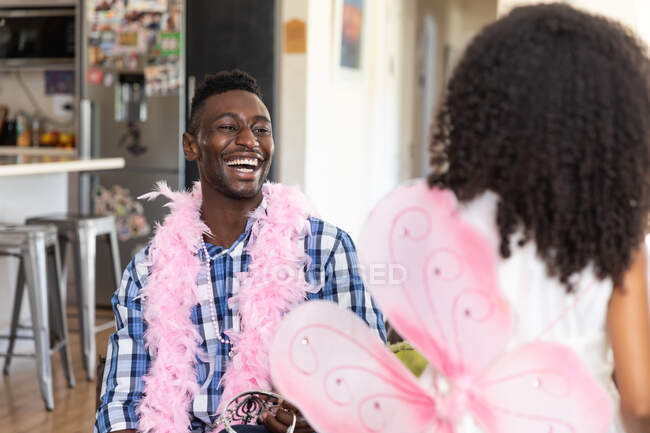Menina afro-americana vestindo um brinquedo asas de fada, o distanciamento social em casa durante o bloqueio de quarentena, brincando com seu pai vestindo rosa boa, em uma sala de estar . — Fotografia de Stock