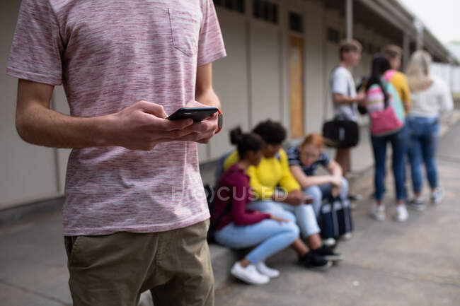 Погляд на середину підлітка з Кавказу, який користується смартфоном, з однокласниками, які висять на задньому плані на шкільному майданчику. — стокове фото