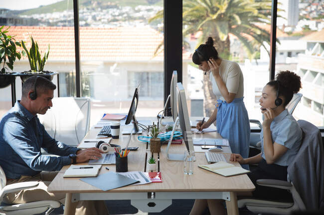 Multiethnische Gruppe männlicher und weiblicher Kollegen, die in einem modernen Büro arbeiten, am Schreibtisch sitzen, Computer benutzen, Headsets tragen und miteinander reden — Stockfoto