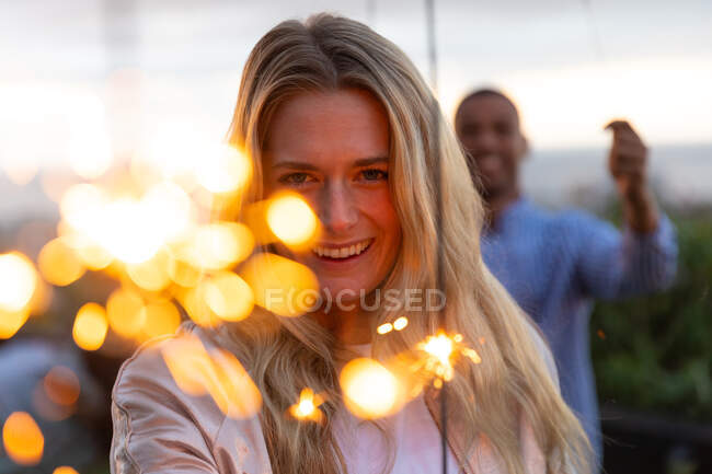 Porträt einer kaukasischen Frau, die auf einer Dachterrasse mit Sonnenuntergang hängt, in die Kamera blickt und lächelt, eine Wunderkerze in der Hand, mit Menschen im Hintergrund — Stockfoto