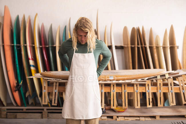 Homem branco surfboard maker em seu estúdio, vestindo um avental de proteção, amarrando atacadores, com pranchas de surf em um rack no fundo . — Fotografia de Stock