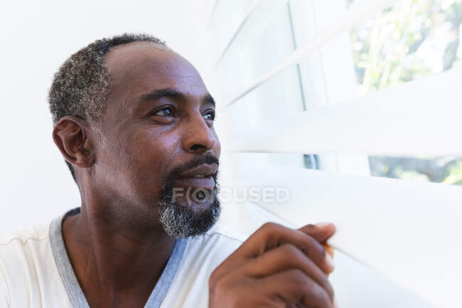Seitliche Nahaufnahme eines gutaussehenden älteren afroamerikanischen Mannes zu Hause, der an einem sonnigen Tag zwischen weißen Fensterläden aus dem Fenster schaut und lächelt, während der Coronavirus-Pandemie selbstisolierend 19 — Stockfoto