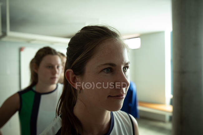 Side view close up of a Caucasian female field hockey player, preparing before a game, standing in a changing room, with her teammates standing in a row behind her — Stock Photo