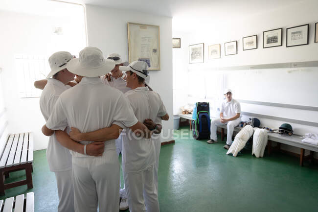 Vue latérale d'un groupe de joueurs de cricket masculins multi-ethniques adolescents portant des blancs, se blottissant dans un vestiaire, avec un autre joueur reposant sur un banc. — Photo de stock