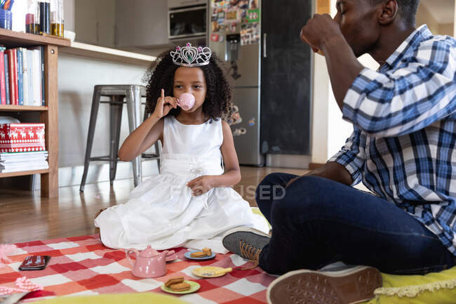 Афроамериканка, одягнена в іграшкову корону, яка дистанціює вдома під час карантину, бавиться з татом, влаштовує лялькову вечірку.. — стокове фото
