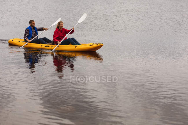 Vista lateral de alto ângulo de um casal caucasiano se divertindo em uma viagem às montanhas, de caiaque em um lago, sorrindo — Fotografia de Stock