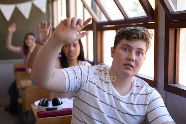 Front view of a teenage Caucasian boy sitting a desk looking ahead and raising his hand in a school classroom, with a row of teenage female classmates sitting at desks behind him also raising their hands — Stock Photo