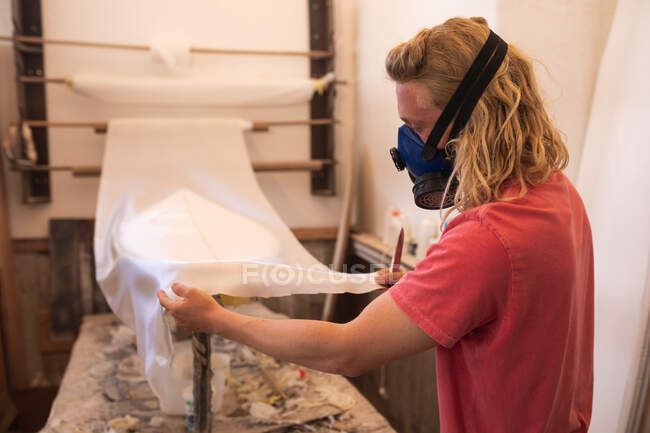 Caucásico fabricante de tablas de surf masculino con una mascarilla respiratoria, trabajando en su estudio, inspeccionando una tabla de surf cubierta con un trozo de tela blanca. - foto de stock