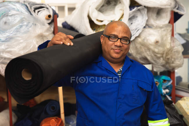 Portrait d'un ouvrier de race mixte dans un atelier d'une usine fabriquant des fauteuils roulants, portant un rouleau de matériel sur son épaule et regardant la caméra — Photo de stock