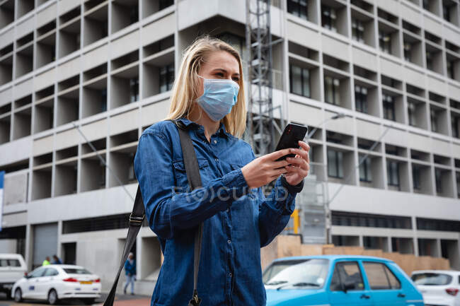 Передній вид жінки, одягненої в маску обличчя проти забруднення повітря і covid19 коронавірусу, яка йде вулицями міста, використовуючи свій смартфон.. — стокове фото
