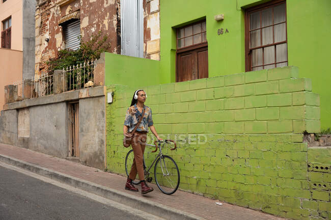 Vista frontal de un hombre de raza mixta con largas rastas en la ciudad en un día soleado, usando auriculares, caminando por la calle y moviendo su bicicleta. - foto de stock
