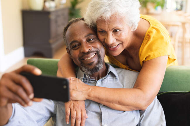 Close up de um casal afroamericano aposentado sênior feliz em casa em sua sala de estar, o homem sentado em um sofá segurando um smartphone a mulher de pé atrás e abraçando-o, olhando para o telefone juntos — Fotografia de Stock
