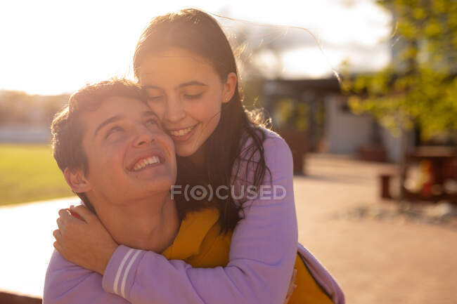 Front close up of a Caucasian teenage girl and boy embracing and smiling at each other standing in the sun in their school grounds — Stock Photo