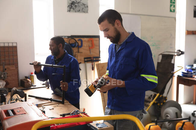 African American and Caucasian male workers in a workshop at a factory making wheelchairs, standing at a workbench and inspecting parts, wearing a workwear — Stock Photo