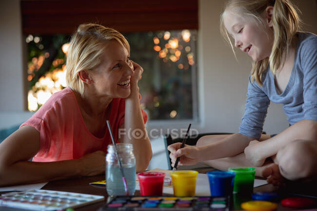 Seitenansicht einer kaukasischen Frau, die Familienzeit mit ihrer Tochter zu Hause genießt, an einem Tisch in einem Wohnzimmer sitzt, malt und lächelt, die Tochter sitzt auf einem Tisch — Stockfoto