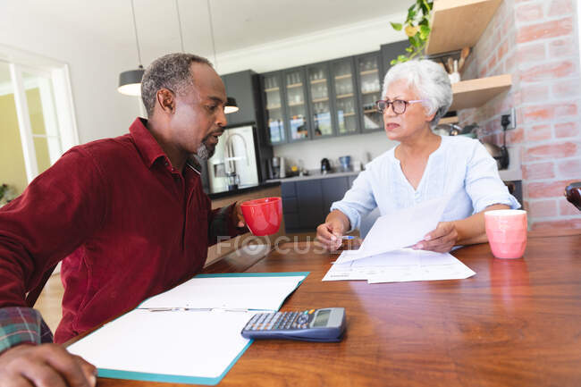 Una pareja de ancianos afroamericanos jubilados sentados en una mesa en su comedor bebiendo café, mirando el papeleo y discutiendo sus finanzas, en casa juntos aislándose durante la pandemia del coronavirus covid19 - foto de stock