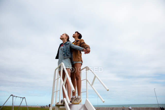 Vista a basso angolo di un caucasico e di una razza mista ragazze godendo di tempo appendere insieme in una giornata di sole, in piedi insieme sulla cima di uno scivolo. — Foto stock
