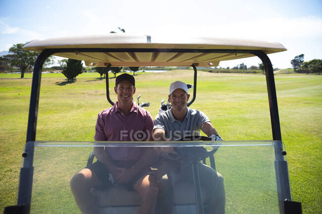 Retrato de dos hombres caucásicos en un campo de golf en un día soleado con cielo azul, conduciendo un carrito de golf, sonriendo a la cámara - foto de stock