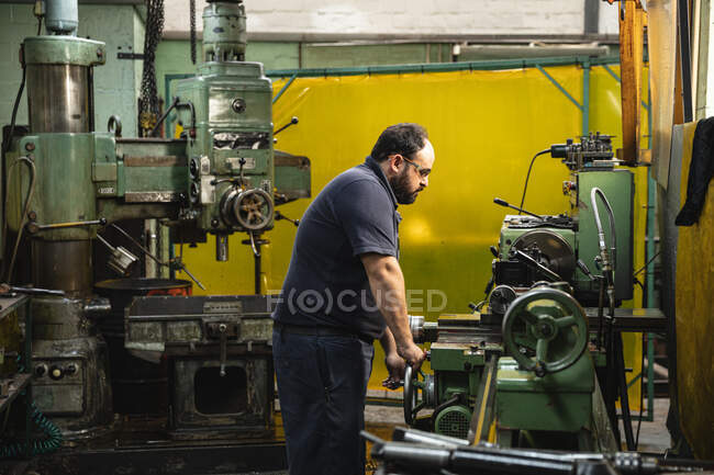 Caucasian male factory worker in a workshop making hydraulic equipment, wearing safety glasses, operating machinery. — Stock Photo