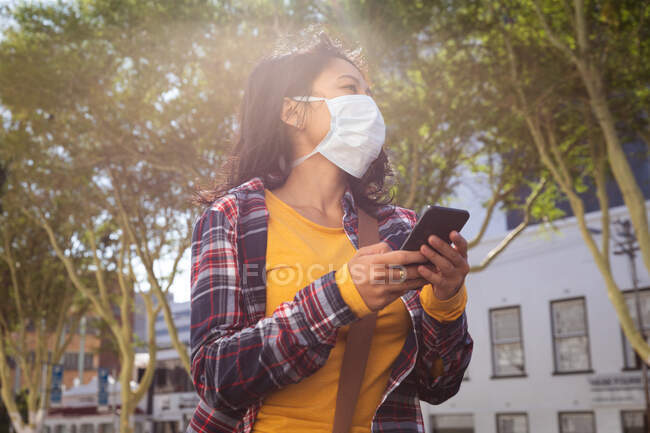 Low angle side view of a mixed race woman with long dark hair out and about in the city streets during the day, wearing a face mask against air pollution and coronavirus, standing and using a smartphone with trees and buildings in the background. — Stock Photo