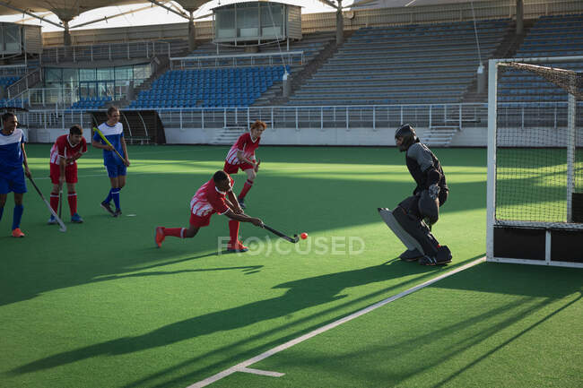 Side view of a teenage mixed race male field hockey player taking a shot at goal during a field hockey match, with player form both teams in the background and the goalkeeper preparing to block the shot, on a sunny day — Stock Photo