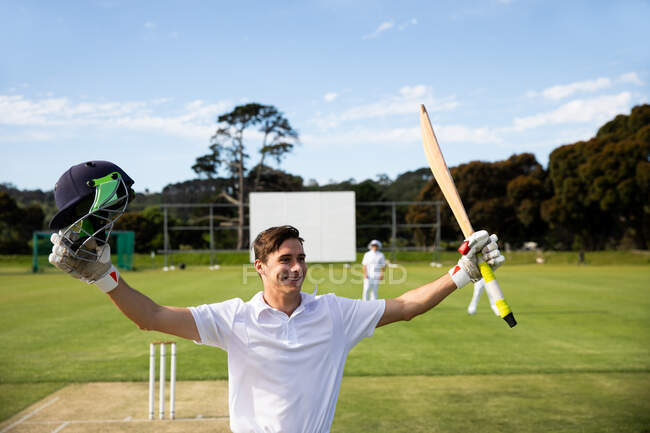 Vorderansicht eines jugendlichen kaukasischen männlichen Cricketspielers, der Weiße trägt, auf dem Spielfeld steht, lächelt und die Hände hebt, einen Cricketschläger und einen Crickethelm in der Hand hält. — Stockfoto