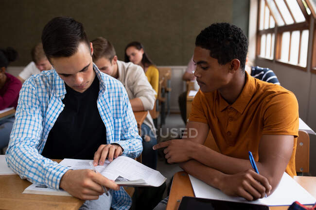 Frontansicht eines kaukasischen Teenagerjungen und eines gemischten Teenagers in einem Klassenzimmer, an einem Schreibtisch sitzend, zusammen arbeitend, in ihren Büchern schreibend und lesend, mit männlichen und weiblichen Teenagern — Stockfoto