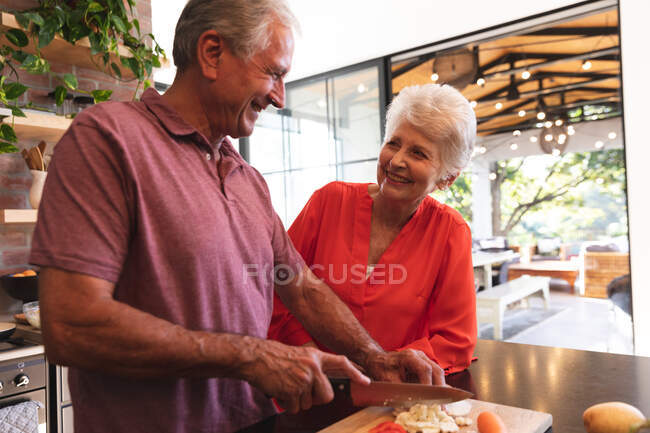 Feliz pareja caucásica jubilada en casa, preparando comida y sonriendo en su cocina, el hombre cortando verduras, la mujer sonriéndole, en casa aislándose durante la pandemia del coronavirus covid19 - foto de stock