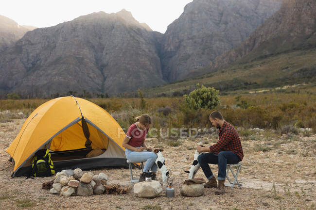 Side view of a Caucasian couple having a good time on a trip to the mountains, sitting by a campfire, playing with puppies — Stock Photo