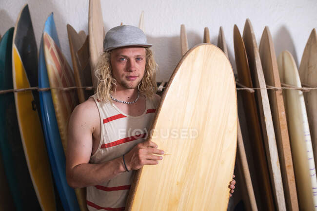 Creatore di tavole da surf caucasiche nel suo studio, che tiene una delle tavole da surf e sorride alla telecamera, con altre tavole da surf in una cremagliera sullo sfondo. — Foto stock