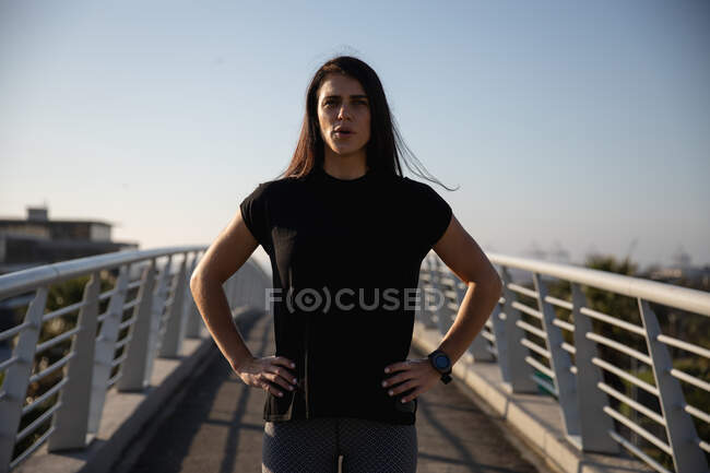 Портрет белой женщины с длинными темными волосами в спортивной одежде на улице в солнечный день с голубым небом, смотрящей на камеру, стоящую на пешеходном мосту — стоковое фото