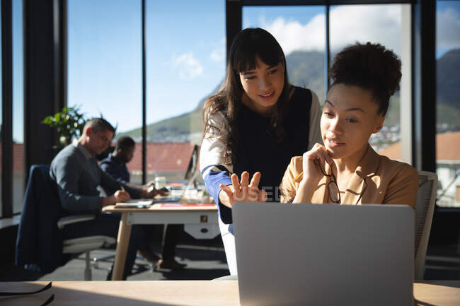 Eine gemischte Rasse und eine asiatische Geschäftsfrau, die in einem modernen Büro arbeitet, einen Laptop benutzt und spricht, während ihre Kollegen im Hintergrund arbeiten — Stockfoto