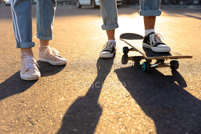 Low section close up of girls enjoying time hanging out together on a sunny day, wearing jeans and trainers, one girl with her foot on the skateboard. — Stock Photo