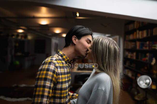 Vista lateral de um jovem homem de raça mista e uma jovem mulher caucasiana desfrutando do tempo em casa, em pé em sua sala de estar e beijando . — Fotografia de Stock
