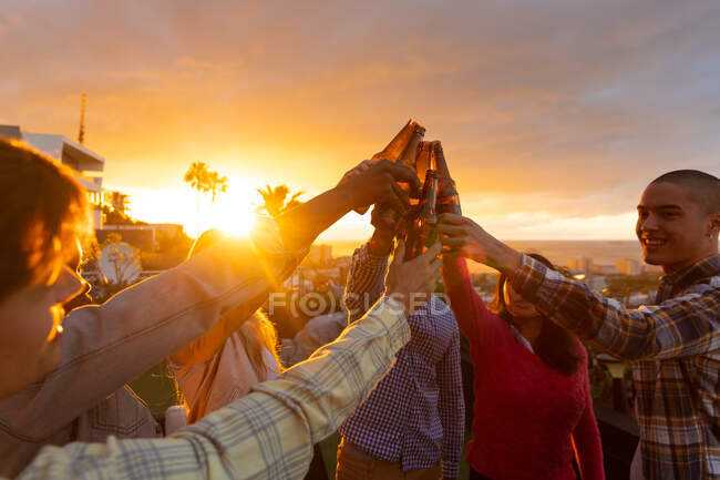 Вид спереди на многонациональную группу друзей, зависающих на террасе на крыше с закатным небом, держащих бутылки пива и произносящих тост — стоковое фото