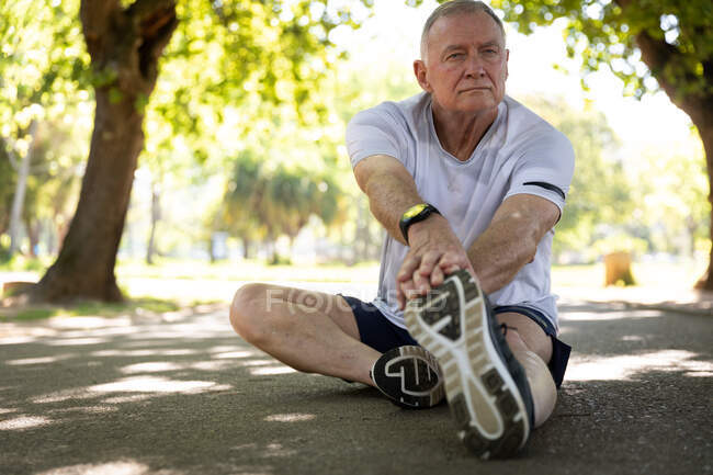 Vista frontale di un maturo uomo caucasico anziano che si allena in un parco in una giornata di sole, seduto sul sentiero e che si estende — Foto stock