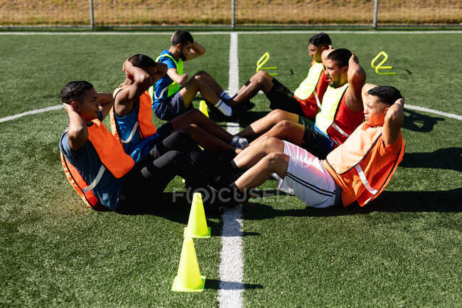 Multi grupo étnico de cinco homens jogadores de futebol um lado vestindo roupas esportivas e coletes de treinamento em um campo de esportes ao sol, aquecendo-se fazendo sentar-se com cones ao lado deles . — Fotografia de Stock