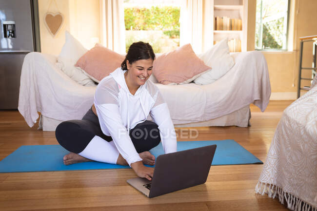 Vlogger femenina caucásica en casa en su sala de estar, practicando yoga y usando su computadora portátil. Distanciamiento social y autoaislamiento en cuarentena. - foto de stock