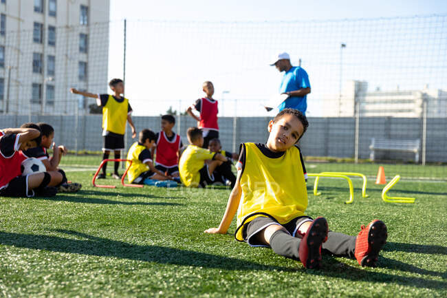 Vue de face d'un footballeur de race mixte assis sur un terrain de jeu au soleil se reposant et regardant vers la caméra pendant une séance d'entraînement, ses coéquipiers écoutant leur entraîneur en arrière-plan — Photo de stock