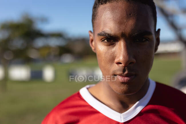 Retrato de um adolescente confiante jogador de rugby masculino mestiço vestindo faixa de equipe vermelha e branca, em pé em um campo de jogo e olhando diretamente para a câmera. — Fotografia de Stock
