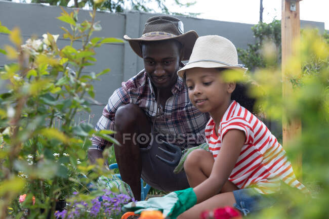 Fille afro-américaine et son père distance sociale à la maison pendant le confinement en quarantaine, passer du temps dans leur jardin ensemble, planter des fleurs, par une journée ensoleillée. — Photo de stock