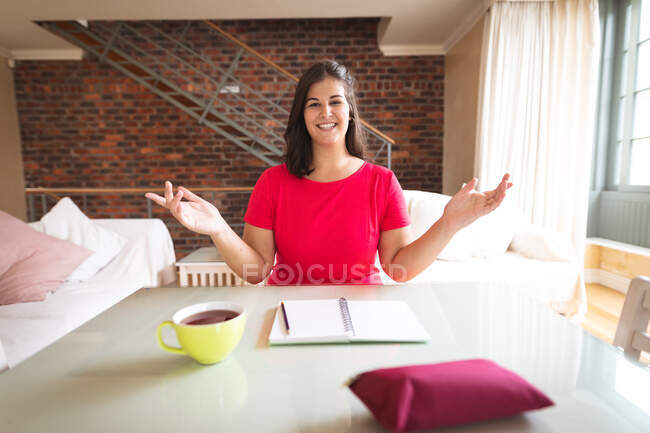 Vlogger mujer caucásica en casa, en su sala de estar preparando su blog en línea, con un cuaderno y una taza de té. Distanciamiento social y autoaislamiento en cuarentena. - foto de stock