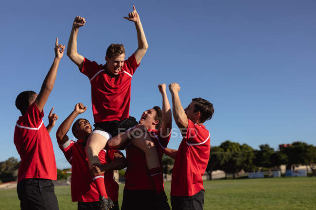 Vista frontal de un grupo de jugadores de rugby masculinos multiétnicos adolescentes que usan tira de equipo roja y blanca, celebrando una victoria, levantando a uno de los jugadores sobre sus hombros y animando con sus brazos en el aire - foto de stock