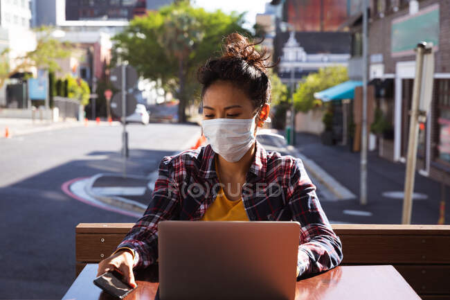 Front view of a mixed race woman with long dark hair sitting at a table in a cafe during the day, wearing a face mask against air pollution and coronavirus, working on a laptop computer using a smartphone with buildings in the background. — Stock Photo