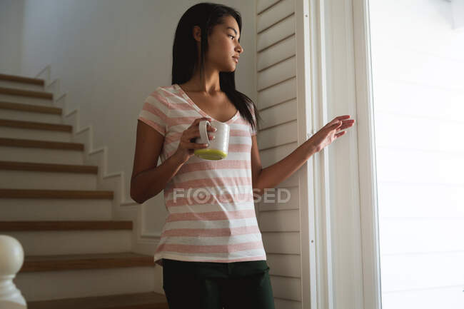 Femme de race mixte passant du temps à la maison, debout et tenant une tasse. Auto-isolement et distanciation sociale en quarantaine pendant l'épidémie de coronavirus covid 19. — Photo de stock