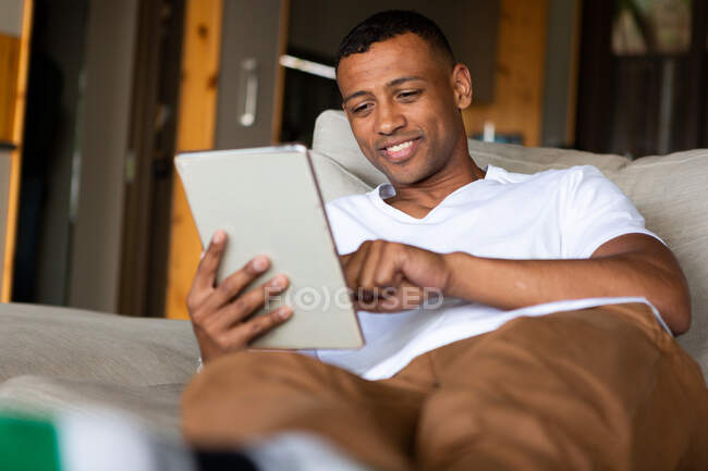 Frontansicht eines afroamerikanischen Mannes, der in seinem Wohnzimmer herumhängt, auf einem Sofa sitzt, einen Laptop benutzt und lächelt — Stockfoto