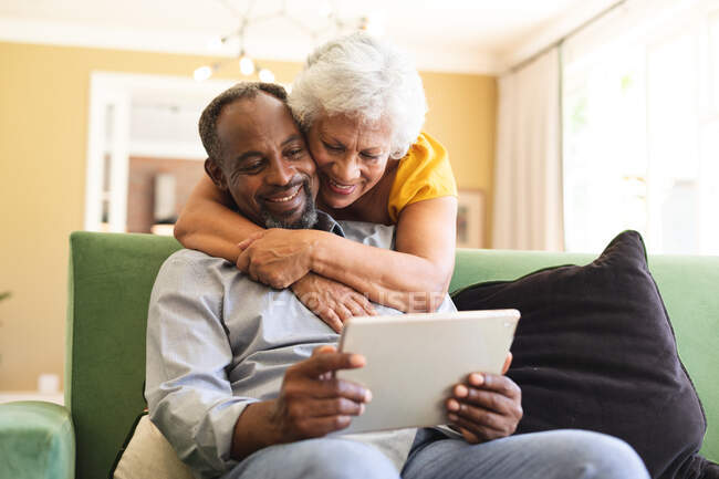 Heureux couple afro-américain retraité à la maison, l'homme assis sur un canapé dans leur salon, la femme debout derrière et l'embrassant, regardant ensemble un ordinateur tablette et souriant, couple isolant pendant la pandémie de coronavirus covid19 — Photo de stock