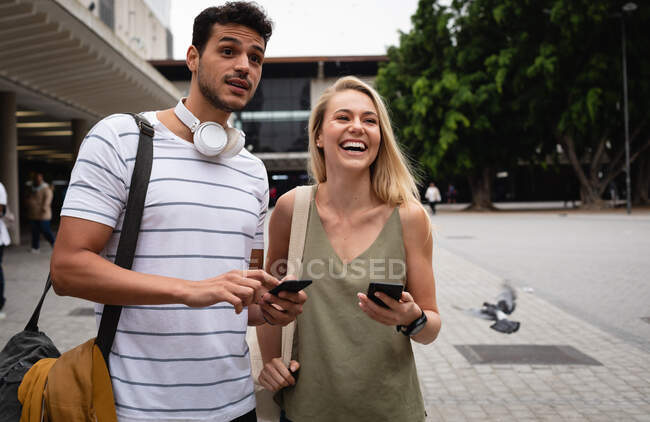 Передній погляд на щасливу кавказьку пару і на вулиці міста протягом дня, стоячи на вулиці і використовуючи свої смартфони.. — стокове фото
