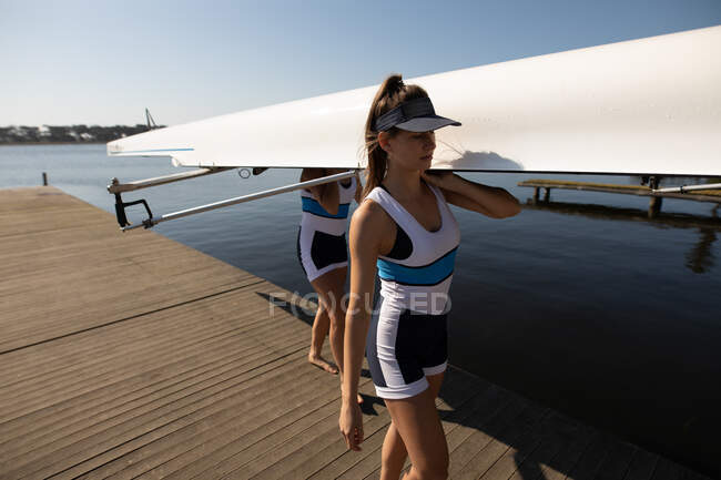 Side view of two Caucasian women from a rowing team training on the river, on a jetty in the sun carrying a boat on their shoulders, the river in the background — Stock Photo