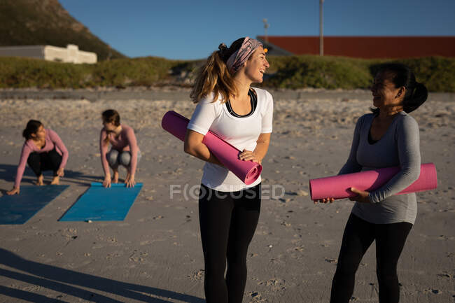 Vista frontale di un gruppo multietnico di amiche che si godono la spiaggia in una giornata di sole, preparando stuoie di yoga per la pratica dello yoga. — Foto stock